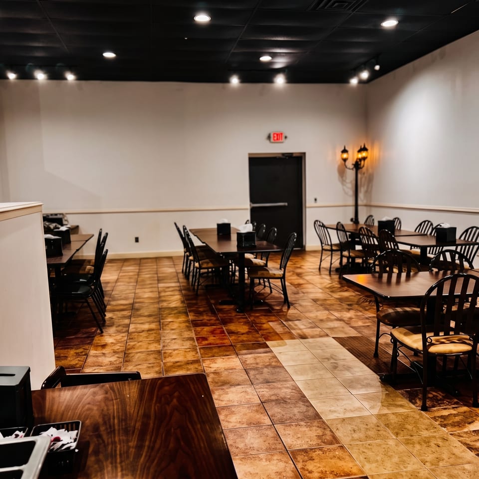 Empty restaurant dining room with dark wood tables, black chairs, tiled floors, warm lighting, and an illuminated exit sign.