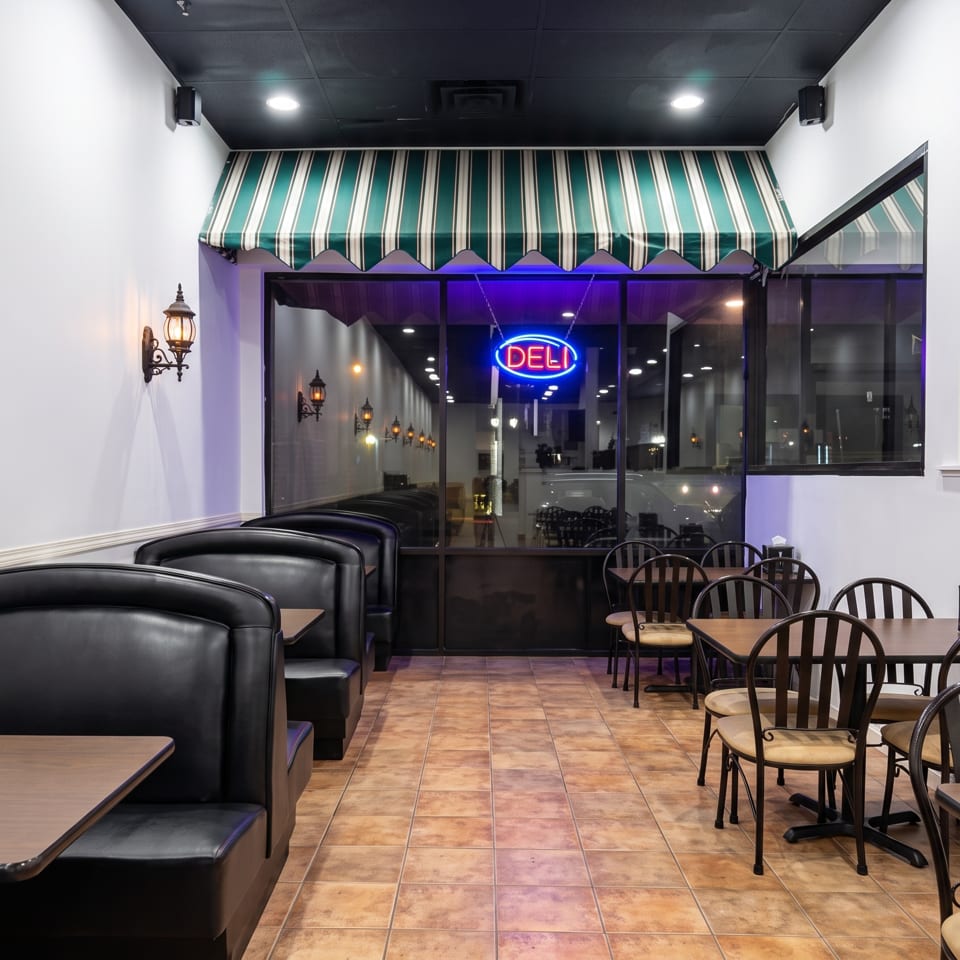 Empty deli dining area with black booths, wooden tables, decorative wall lanterns, tiled floor, and a neon Deli sign.