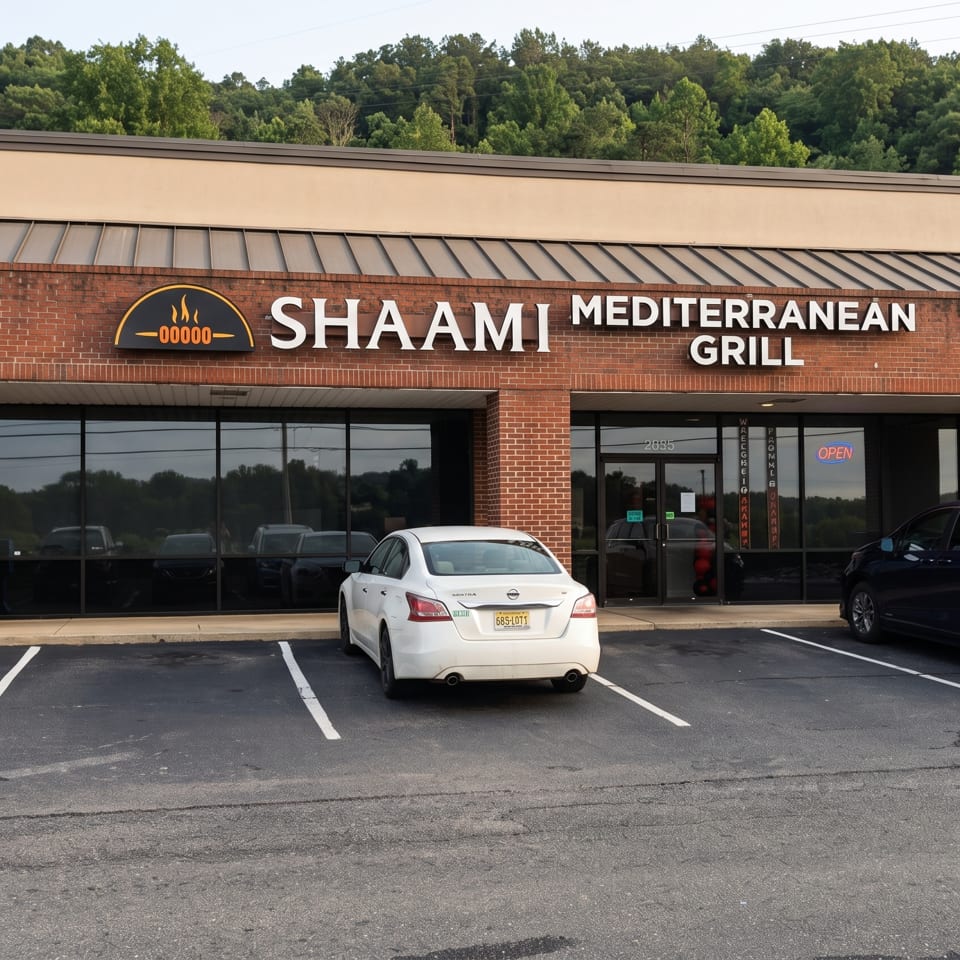 Exterior of Shaami Mediterranean Grill in a brick shopping center, with storefront signage, parked cars, and wooded hills behind.